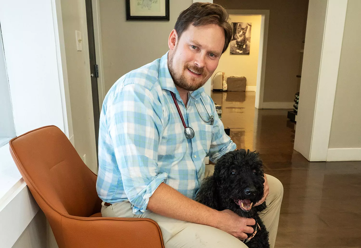 A man in a checkered shirt and beige pants, wearing a stethoscope, sits on a brown chair while holding a black dog in a room with framed pictures and a brown floor.