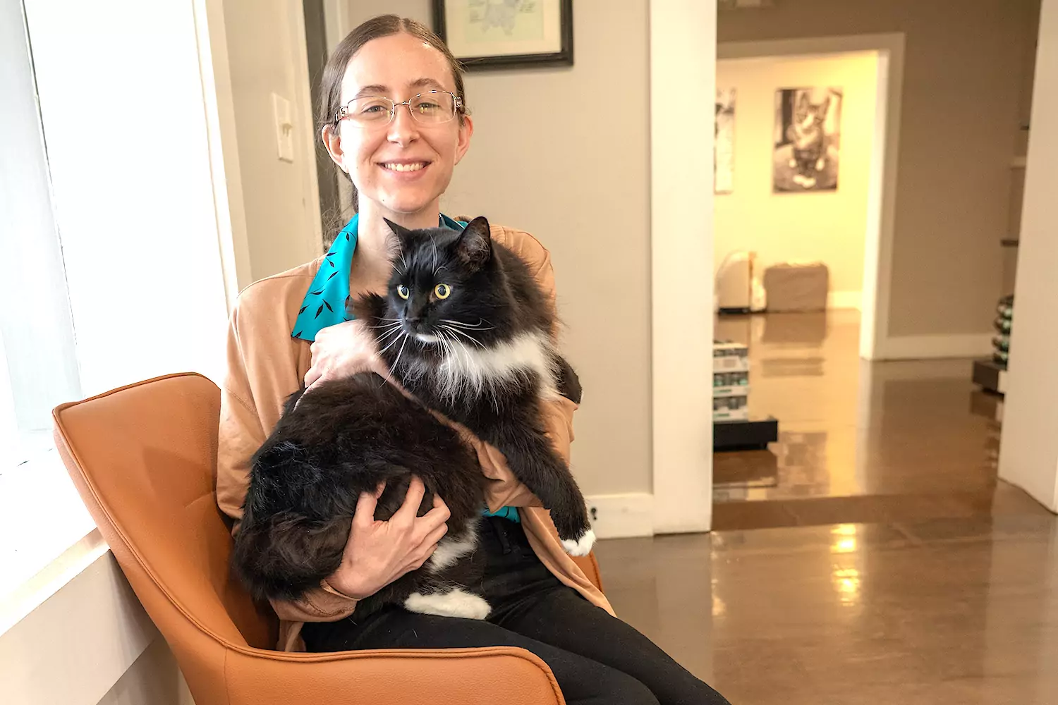 A person with glasses, wearing a brown sweater and a blue shirt, is sitting and holding a large black and white cat indoors.