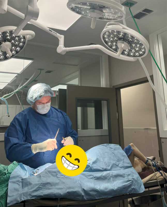 A surgeon in blue scrubs and a mask operates under bright lights in an operating room. A patient lies on the table covered with blue drapes.